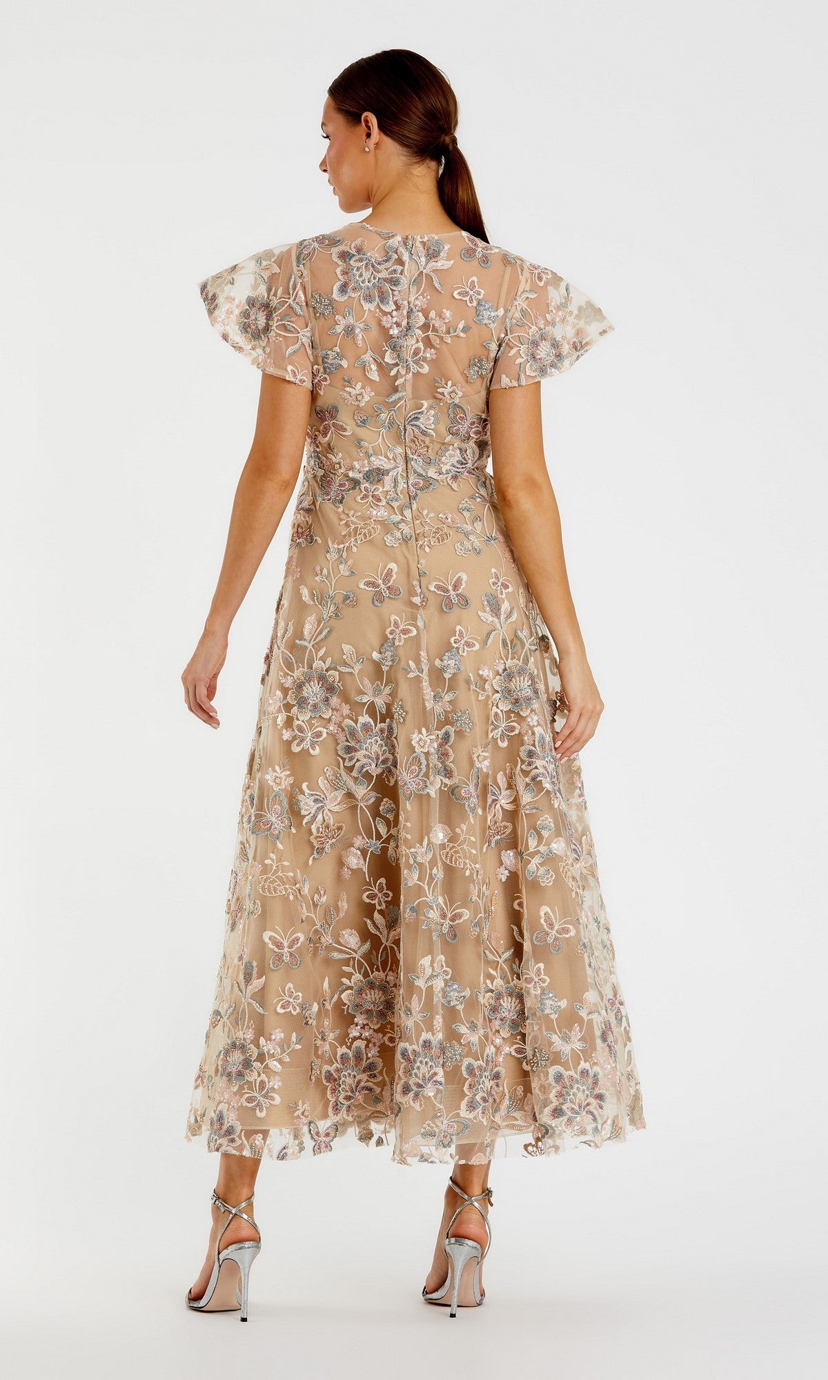 A woman faces away, wearing the Mac Duggal 68920 Tea-Length Party Dress—a sheer beige cocktail dress with floral and beaded embroidery, short flutter sleeves—and silver high heels, standing against a plain white background.